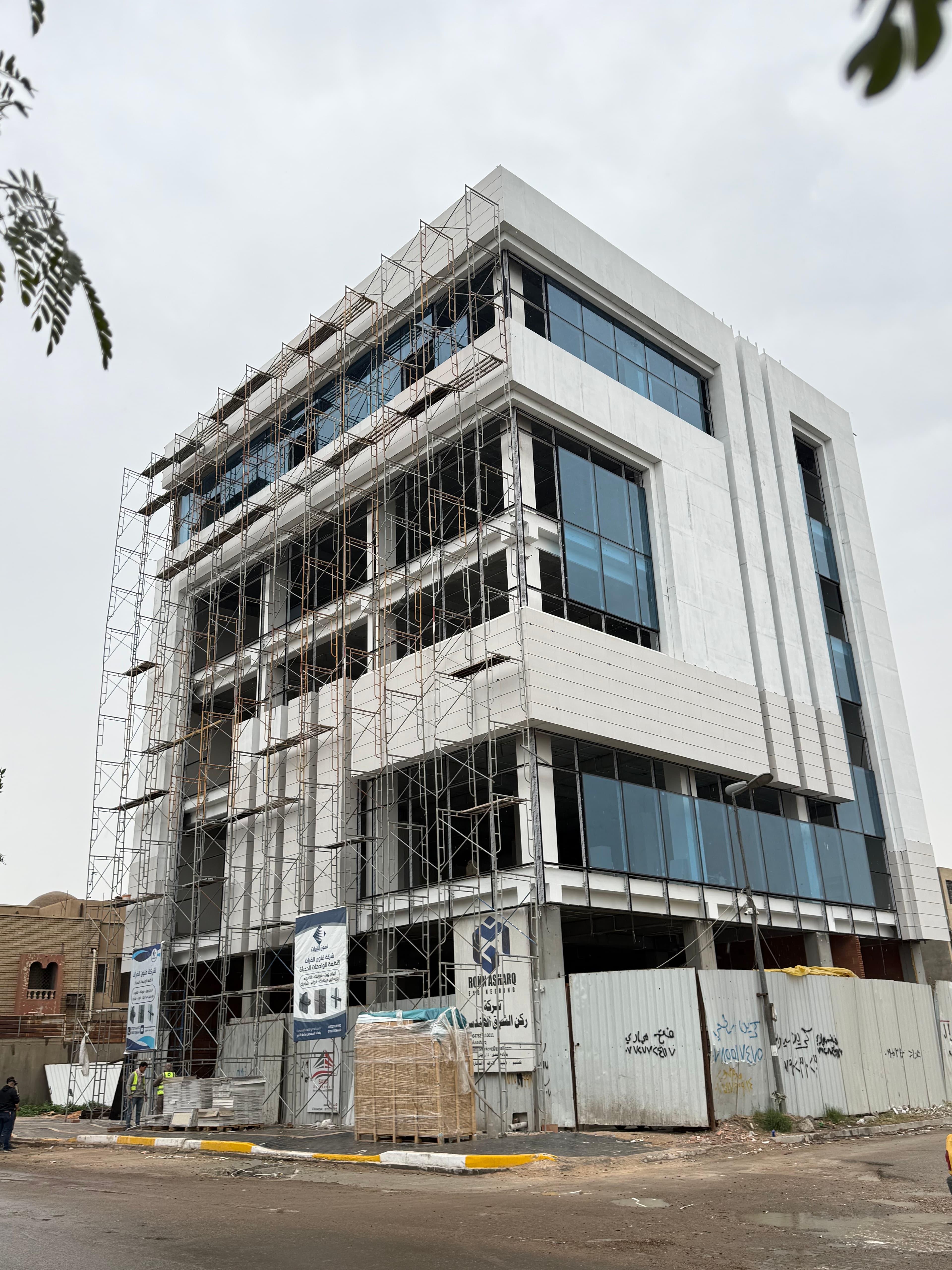 مبنى حديث قيد الإنشاء بواجهة بيضاء ونوافذ زرقاء وسقالات معدنية تحت سماء غائمة.
