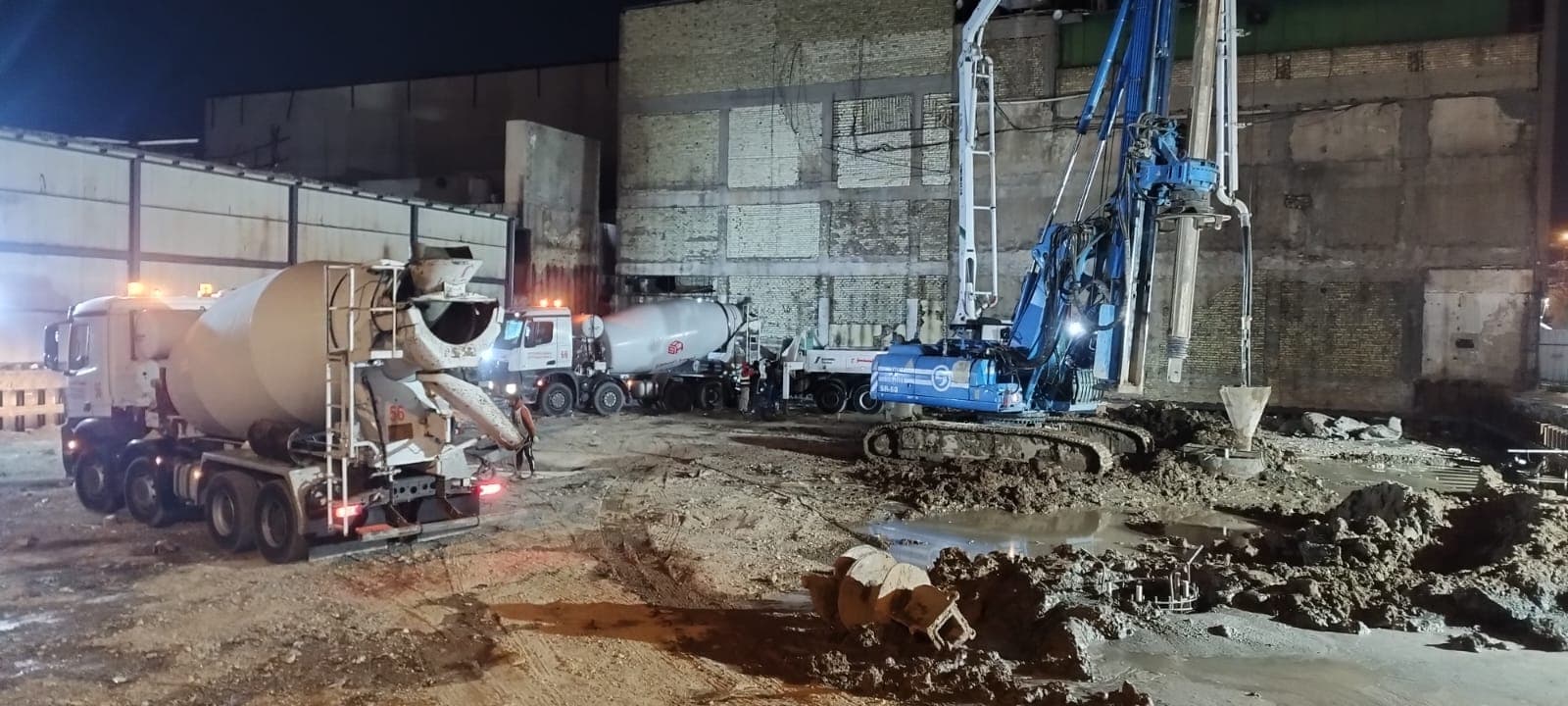 شاحنات خرسانة بيضاء وحفارة زرقاء ضخمة في موقع بناء ليلي مليء بالطين.